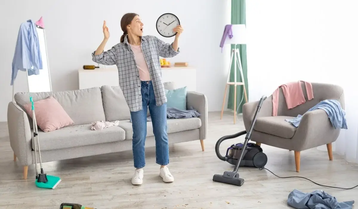 Woman holding clock in cluttered apartment living room with vacuum, timing cleaning